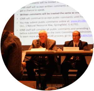 Two people in professional dress at a hearing listening to public questions and preparing to answer