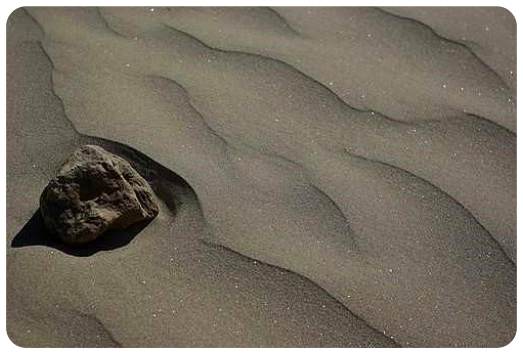 A craggy-textured rock is on the rippled sandy shore of a beach